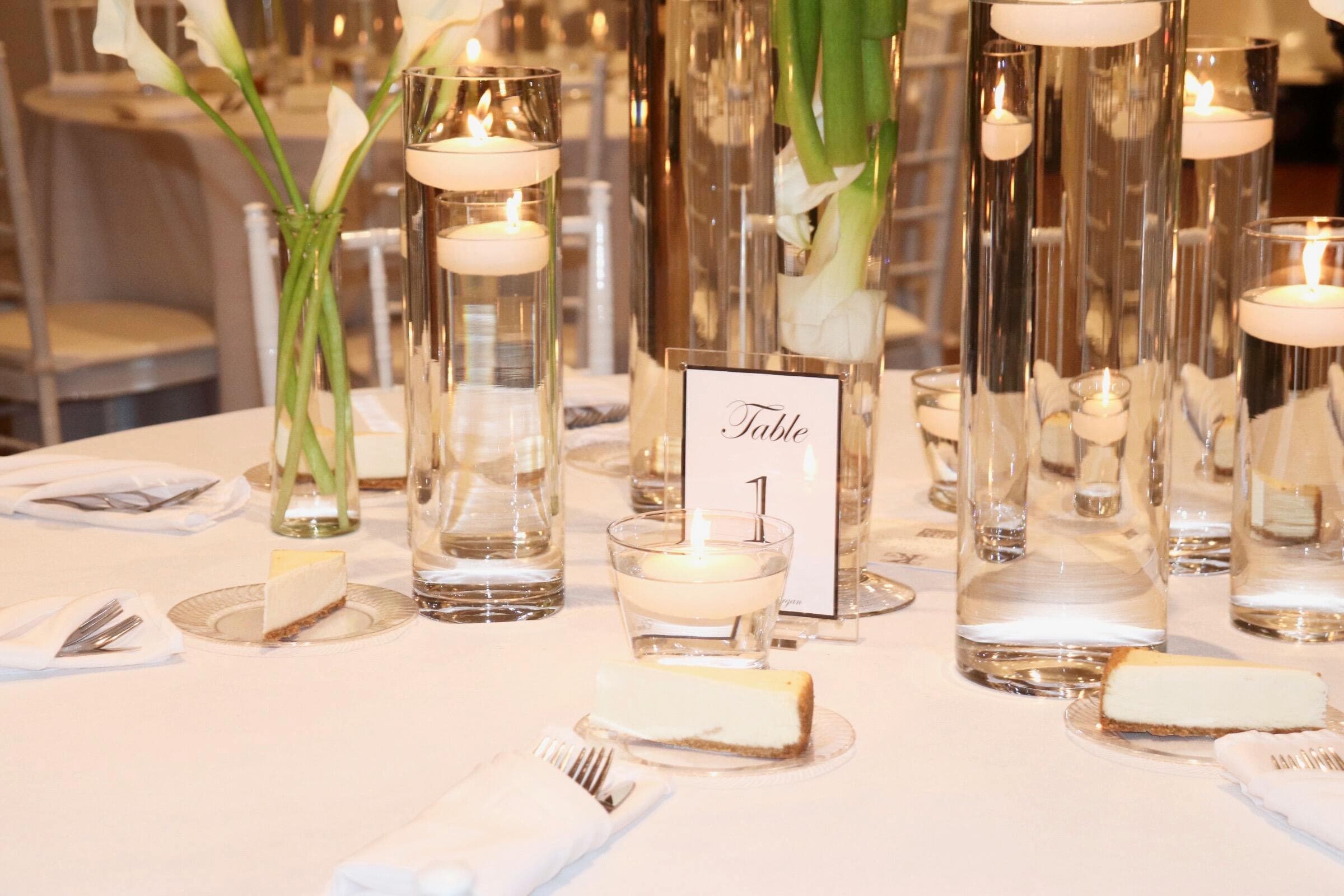 White-clothed reception table featuring tall glass cylinders with floating candles, white calla lilies, a Table 1 sign, and slices of cheesecake