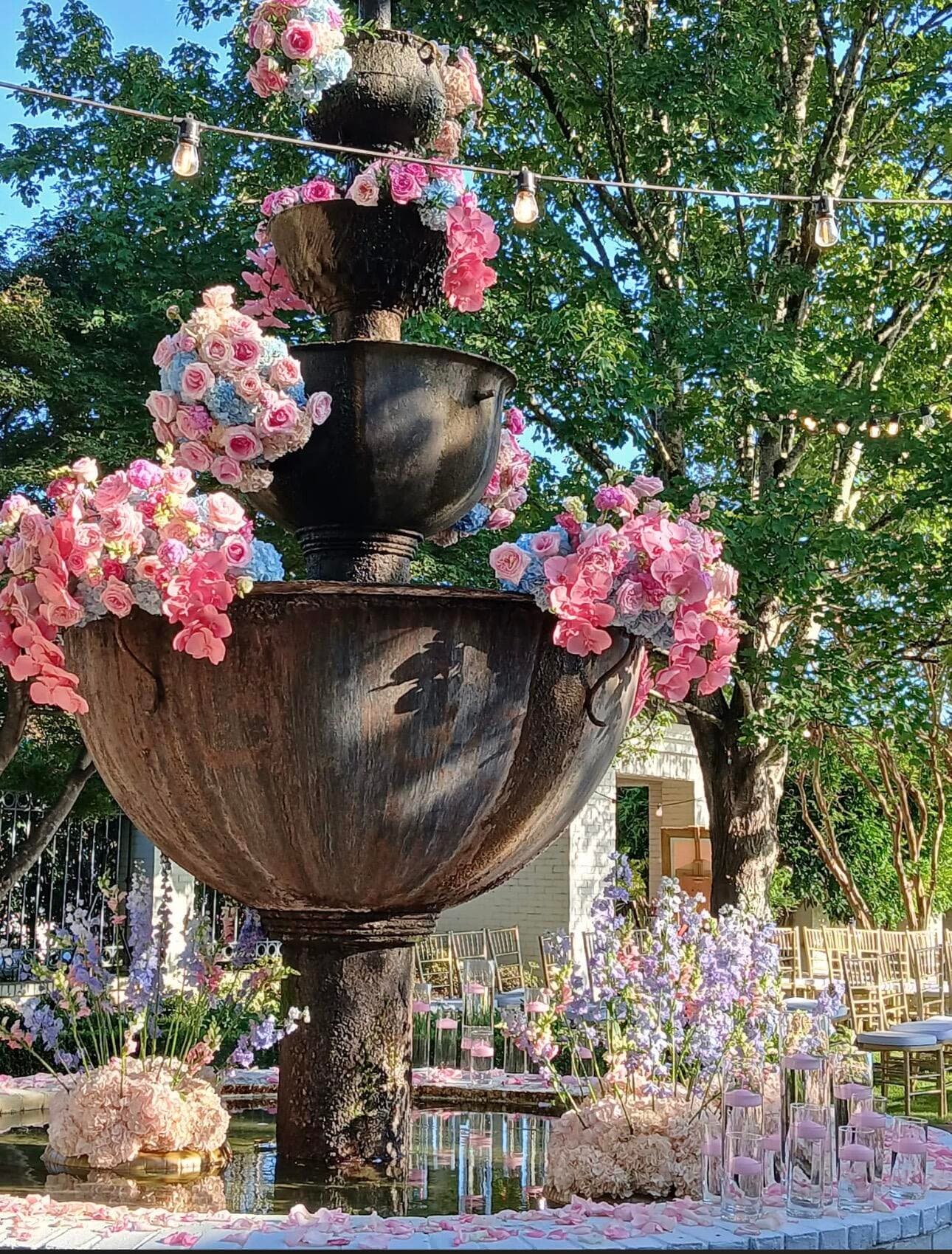Multi-tiered dark stone fountain decorated with pink, blue, and purple floral arrangements and surrounding string lights