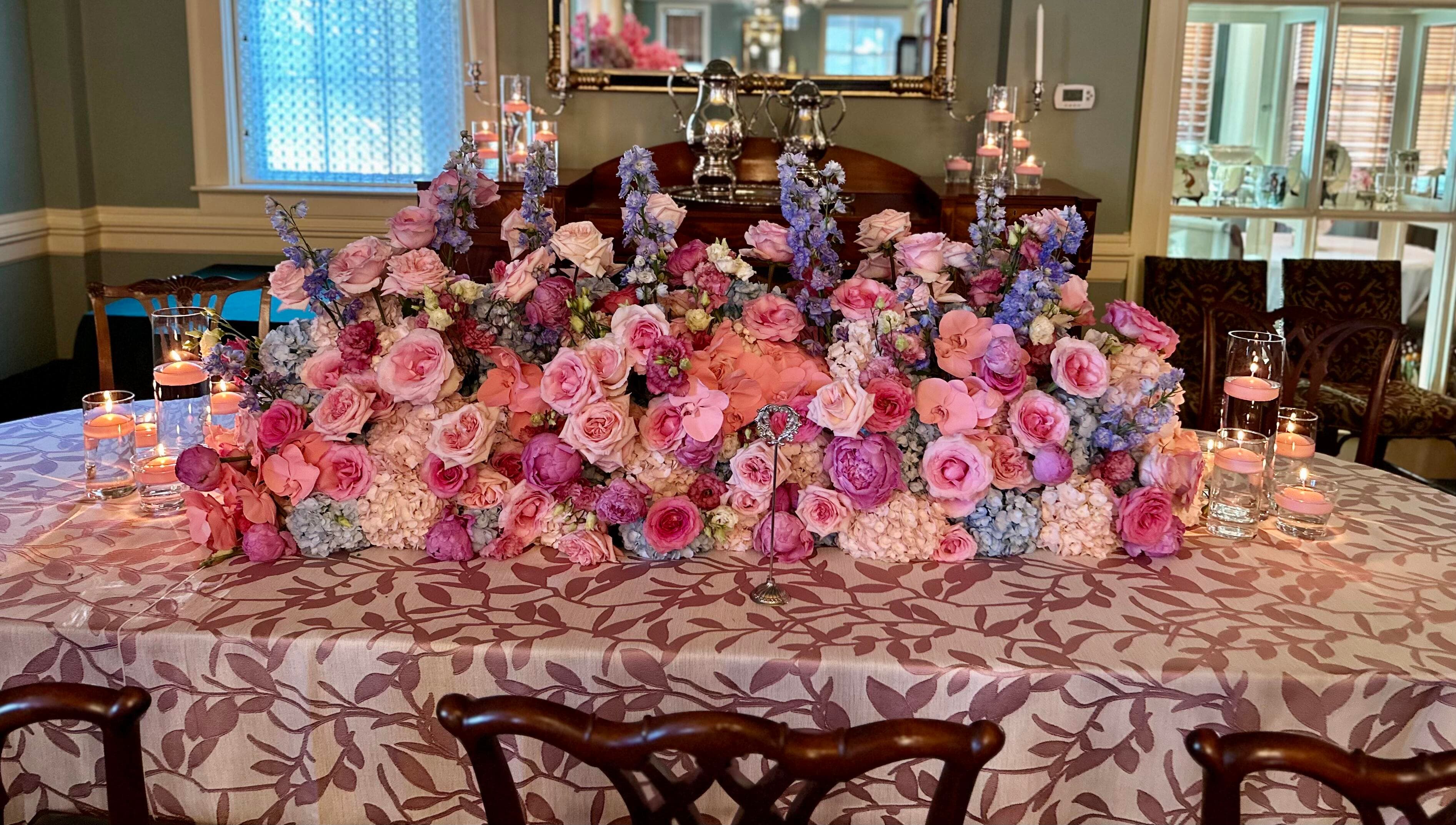 A lush, continuous floral centerpiece runner of pink and purple roses and blue delphiniums down a table with patterned pink linens and glowing votive candles