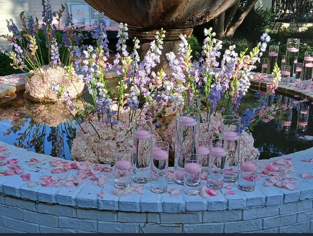 The base of a stone fountain pool filled with pink hydrangeas, purple delphiniums, and floating pink candles in glass cylinders