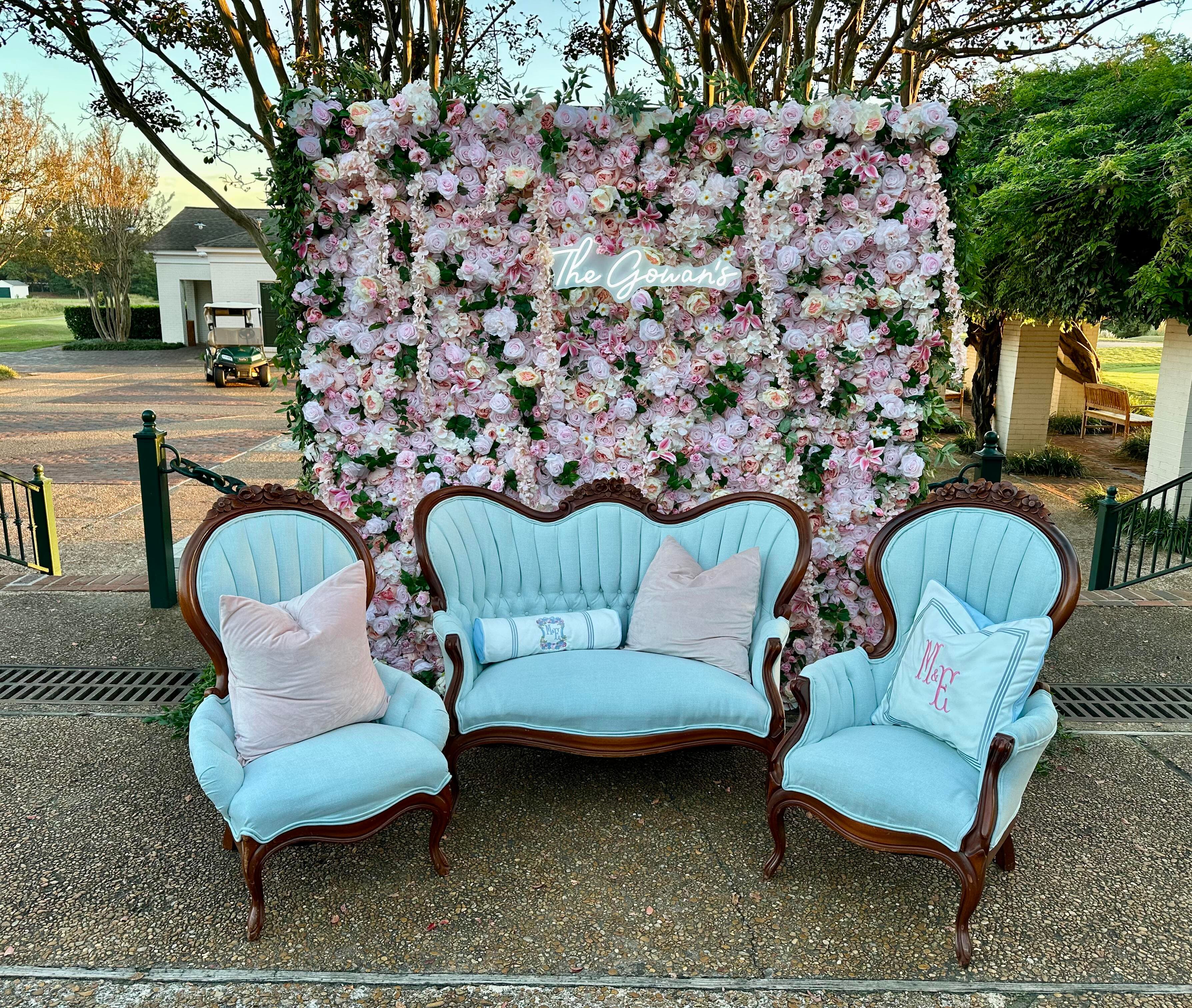 A light blue vintage tufted settee and matching armchairs with custom monogrammed pillows in front of a massive pink and white floral wall backdrop featuring a neon sign