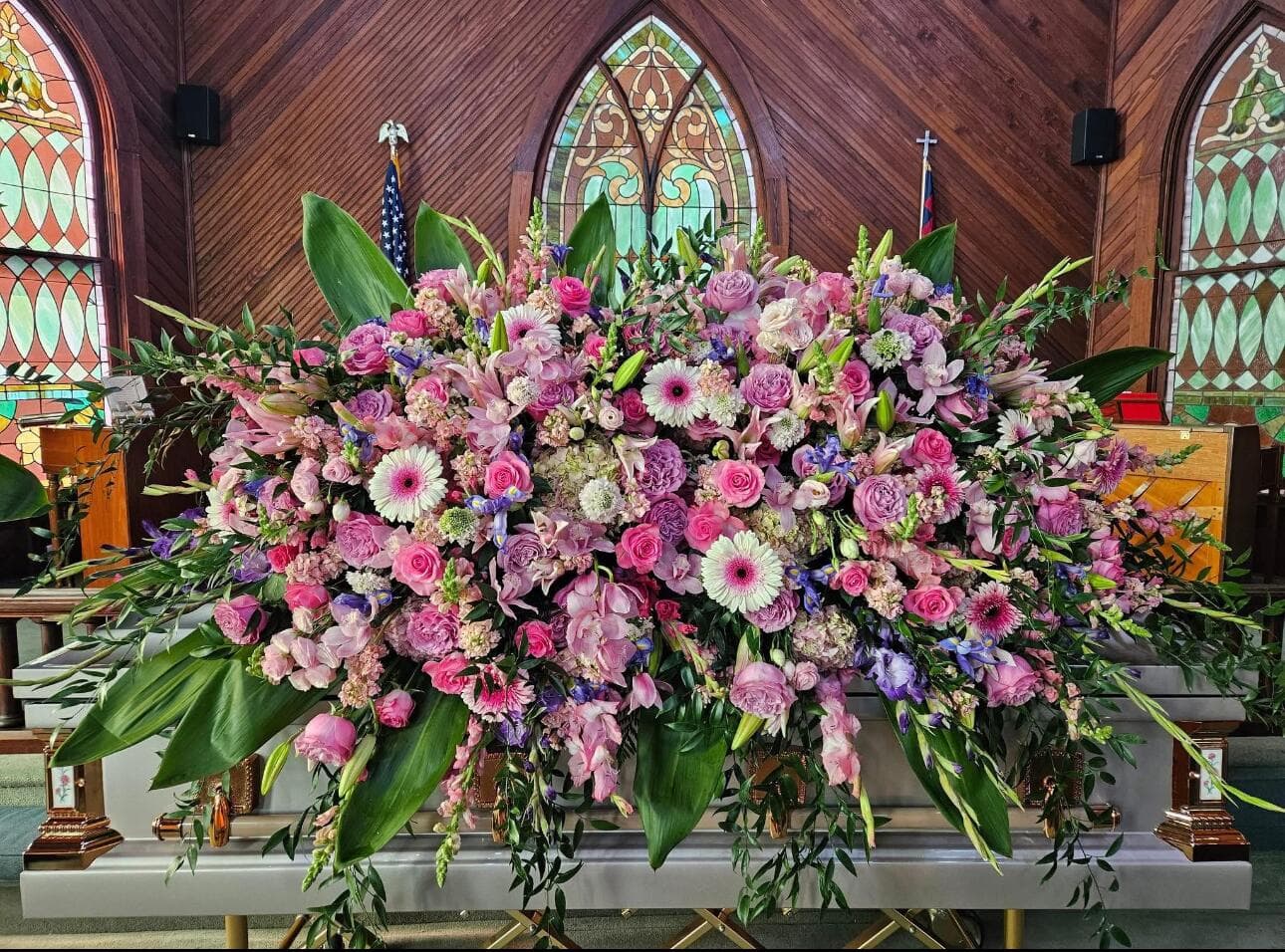 A large casket floral spray featuring a rich mix of pink roses, gerbera daisies, purple accents, and large green tropical leaves inside a wooden church with stained glass windows