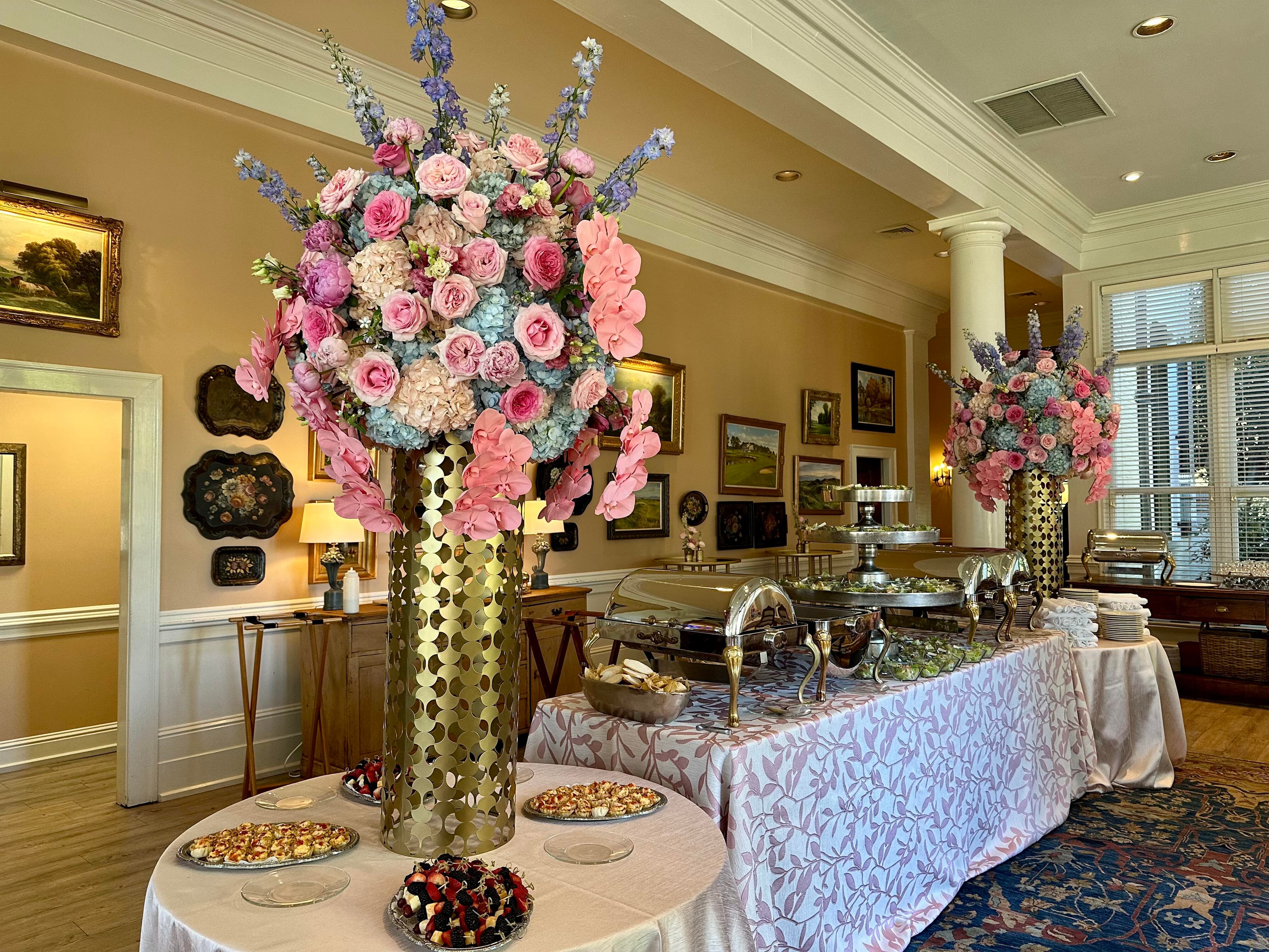 Tall gold patterned cylinder vases holding massive arrangements of pink and blue blooms, set in a banquet hall next to buffet tables with pastries and chafing dishes